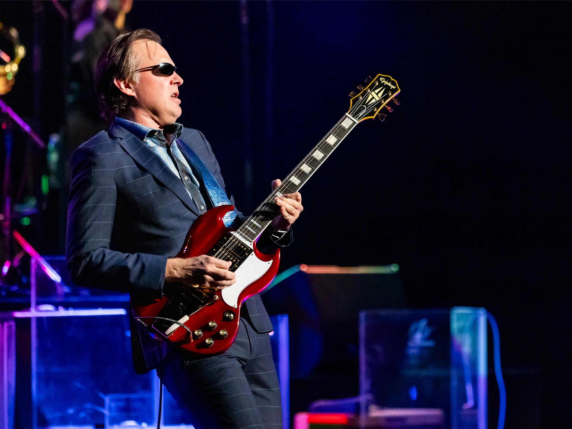 Joe Bonamassa playing guitar on stage. He is wearing a suit and sunglasses and is leaning back as he plays with expression.