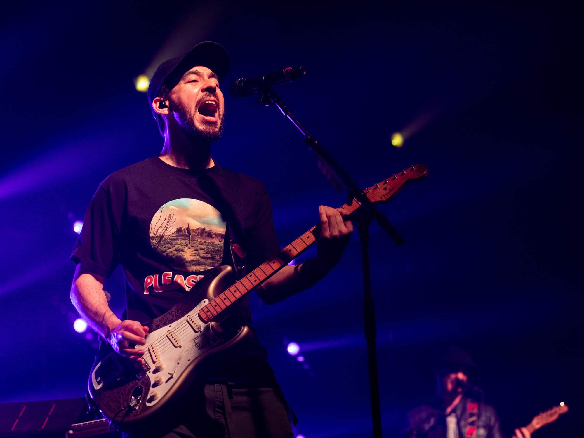 Mike Shinoda on stage. He is playing a Strat and is singing into a mic. He is wearing a dark coloured cap and t-shirt.