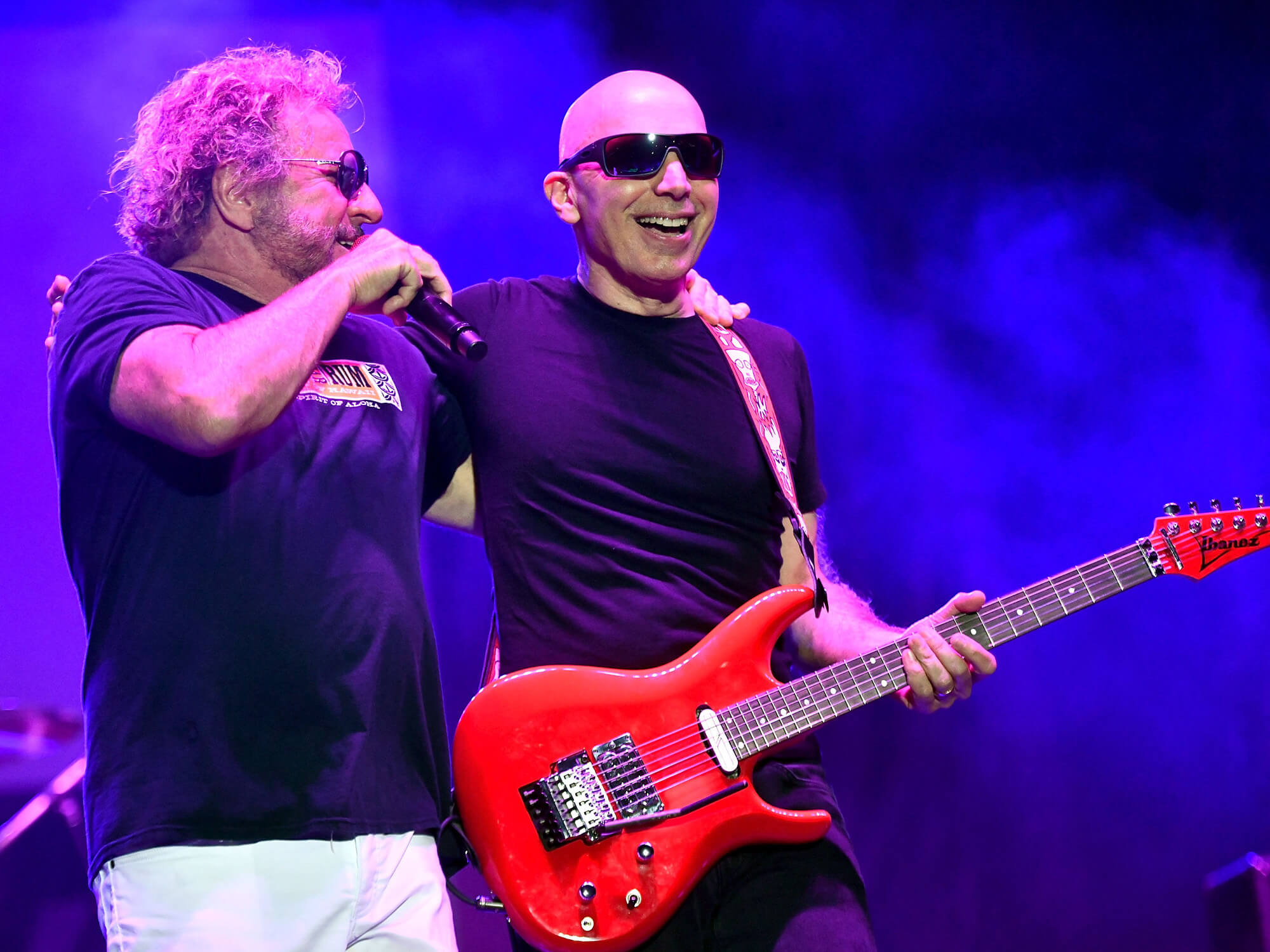 Sammy Hagar with a microphone in one hand, and one arm around Joe Satriani. The pair are on stage, and Satriani is holding a vibrant Ibanez electric model. Both are smiling.