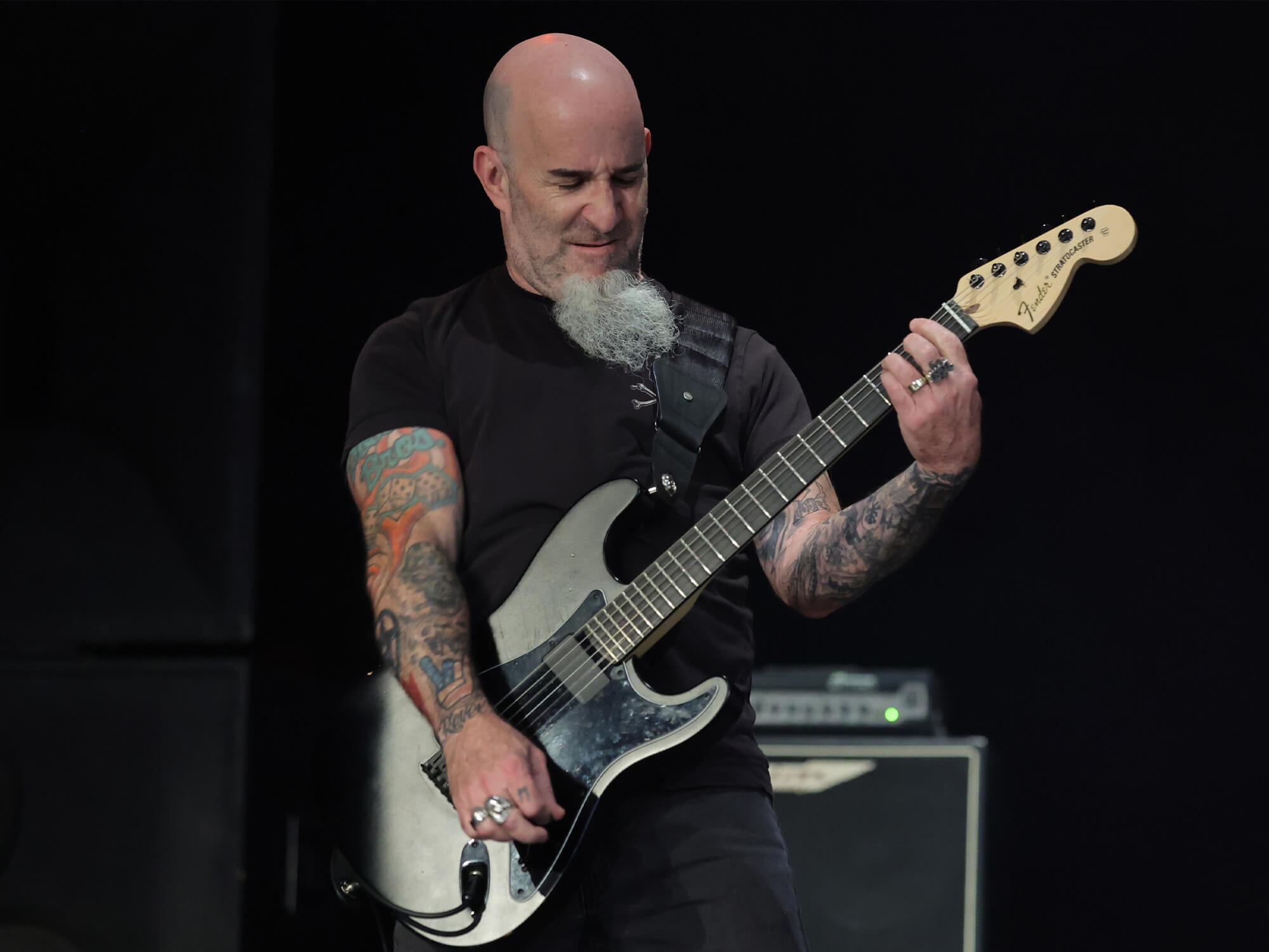 Scott Ian playing a grey Strat guitar. His arm is extended in a large strum and his face is screwed up with passion. He has a long grey beard which runs down from his chin.