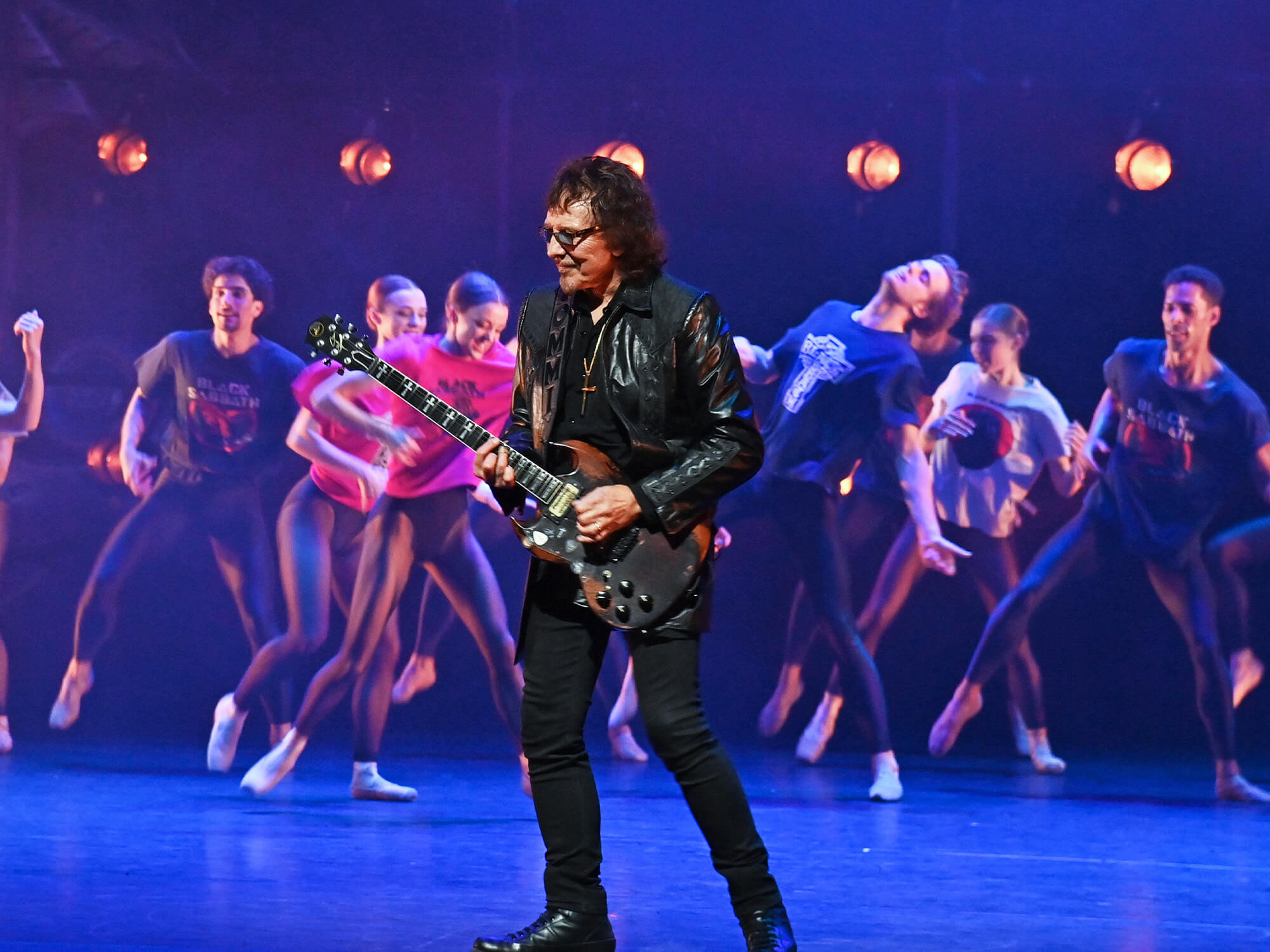 Tony Iommi playing guitar with the Black Sabbath Ballet dancers featured behind him.