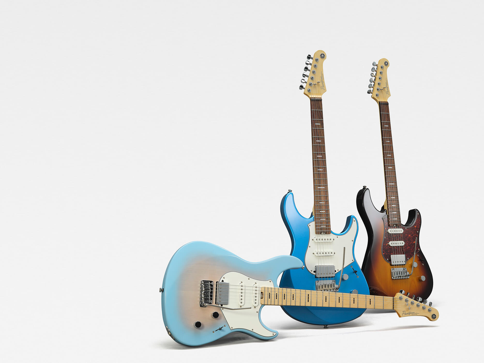 Three new Yamaha Pacifica models. Two are stood upright - one is blue and the other is a warm wooden tone - and a pale blue model with a gradient pink finish is on its side.