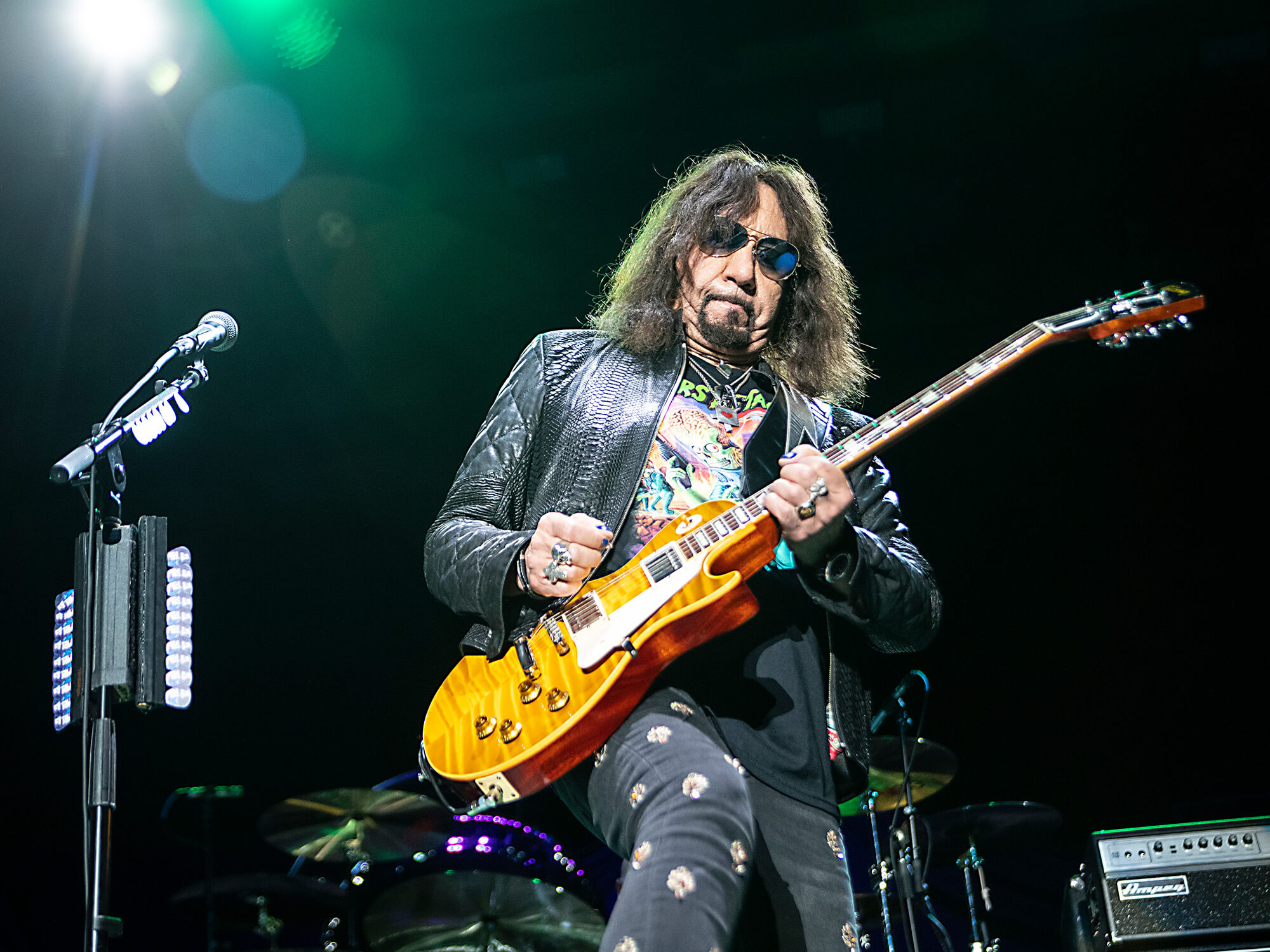 Ace Frehley on stage. The camera is pointing up at him and he is playing guitar. He's wearing shades and dark clothing.