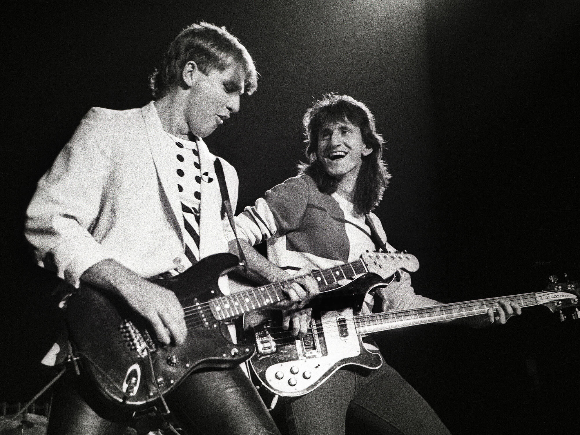 A black and white photo of Alex Lifeson and Geddy Lee on stage in 1983. They both have guitars in hand. Geddy is smiling at Alex, who is looking down at his guitar with a passionate expression. Photo by Rob Verhorst/Getty Images