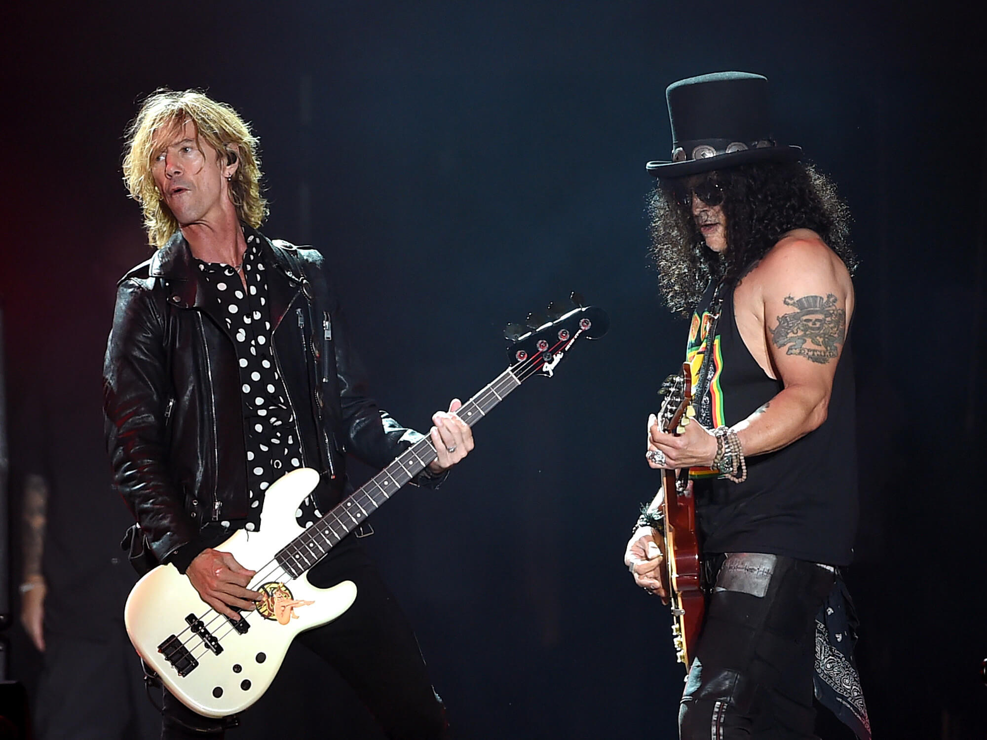 Duff McKagan and Slash on stage. They are turned towards each other. Duff is looking over his shoulder and Slash is looking down. They both have their guitars in hand.