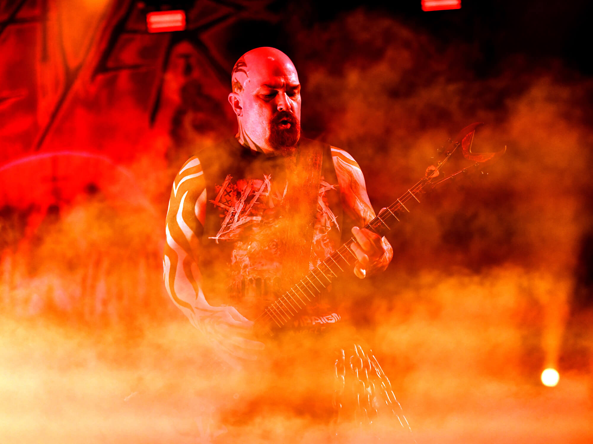Kerry King playing guitar. He is standing among lots of smoke and red lighting.