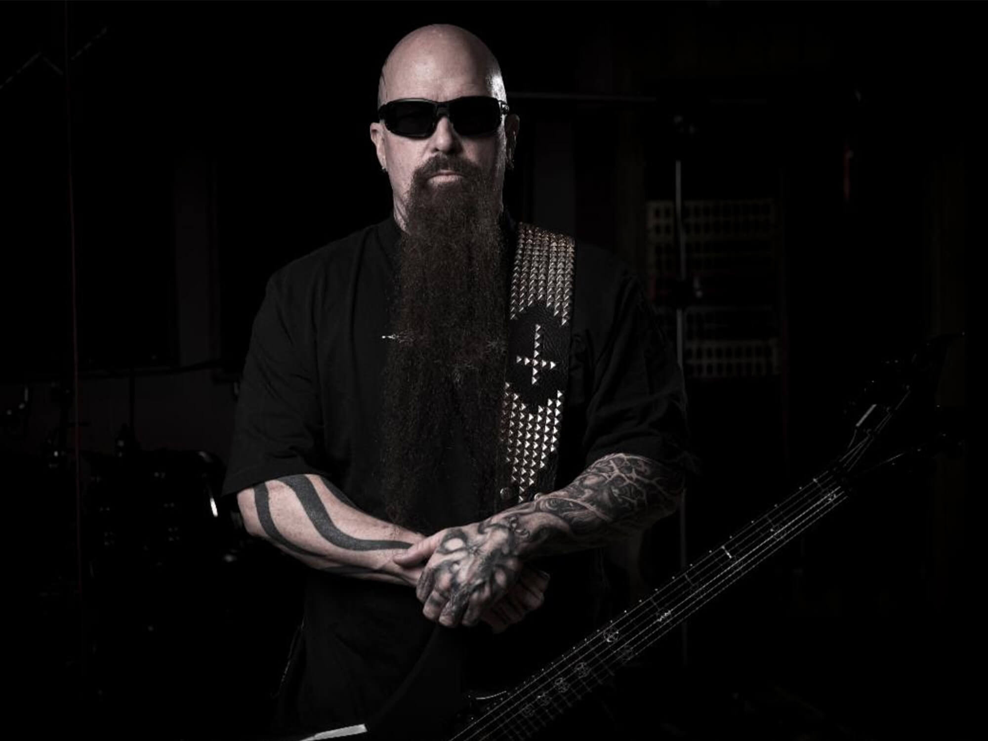 Kerry King standing with a straight expression. He wears all black and stands in front of a black backdrop. He has on shades and wears his guitar on a strap.
