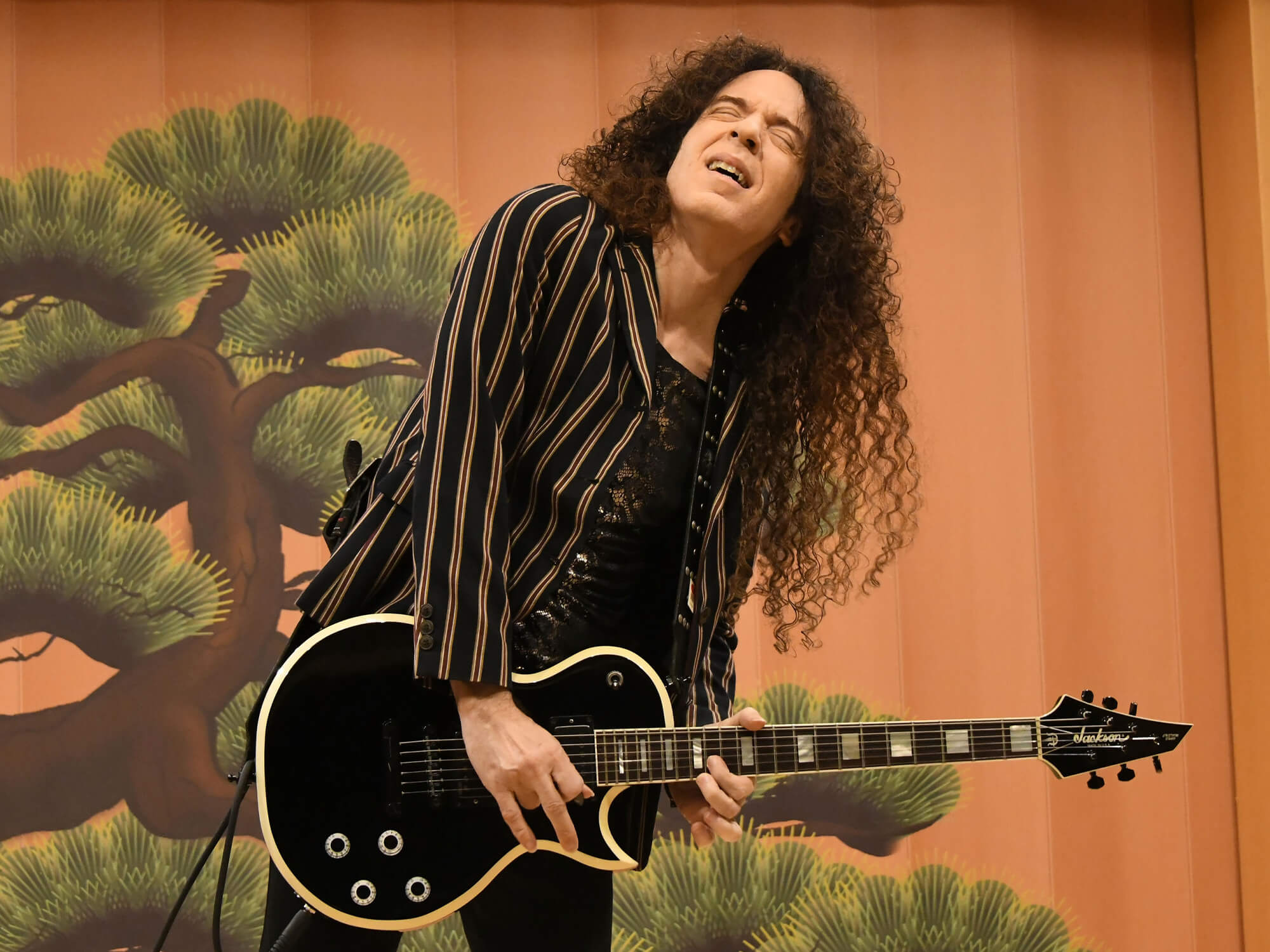 Marty playing guitar. He has his eyes closed and one hand on the high frets, with the other picking the strings. He has long hair and wears dark clothes.
