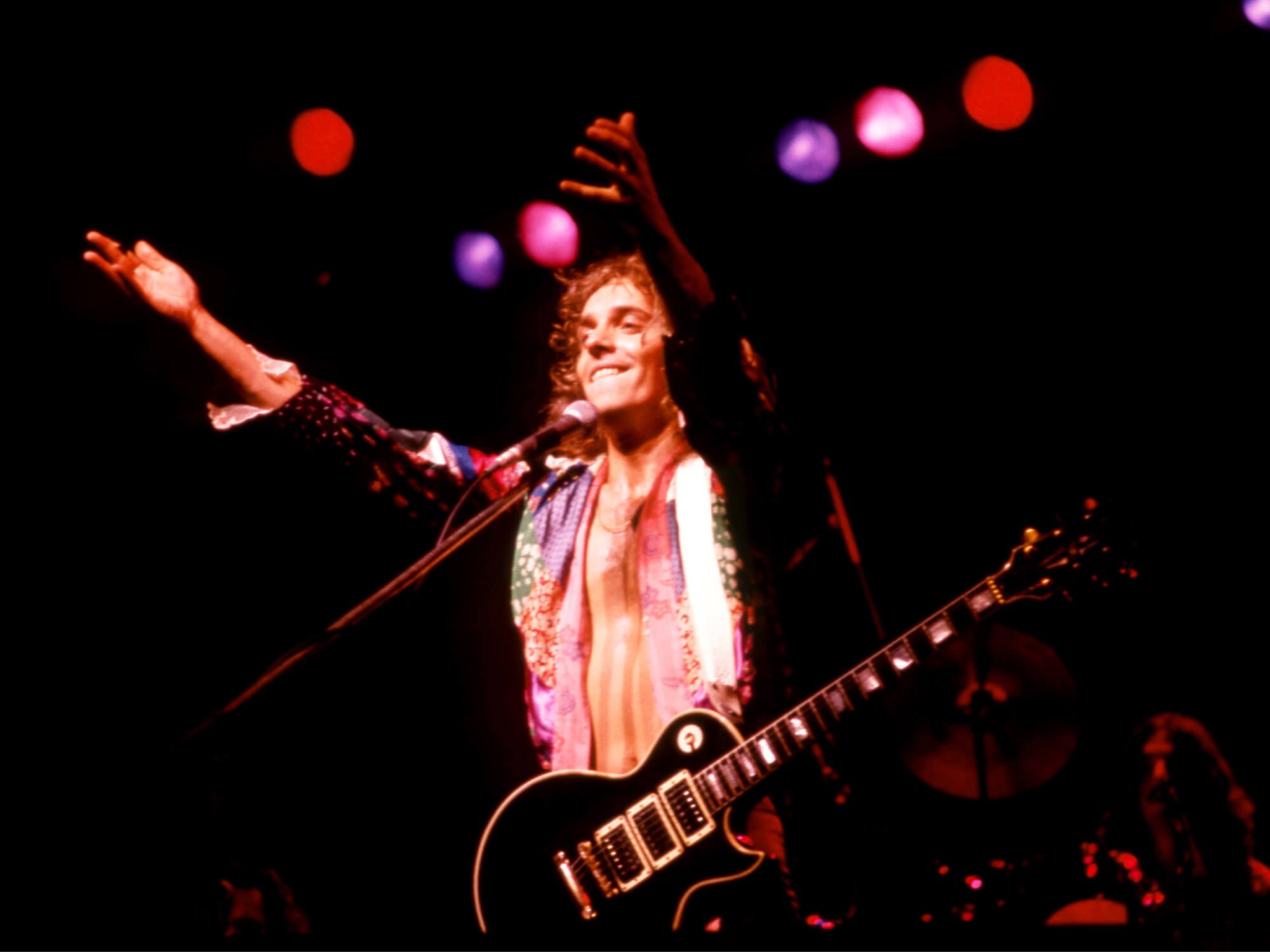 Peter Frampton on stage for his I'm In You Tour. He is holding both arms out in the air towards the audience and is smiling.