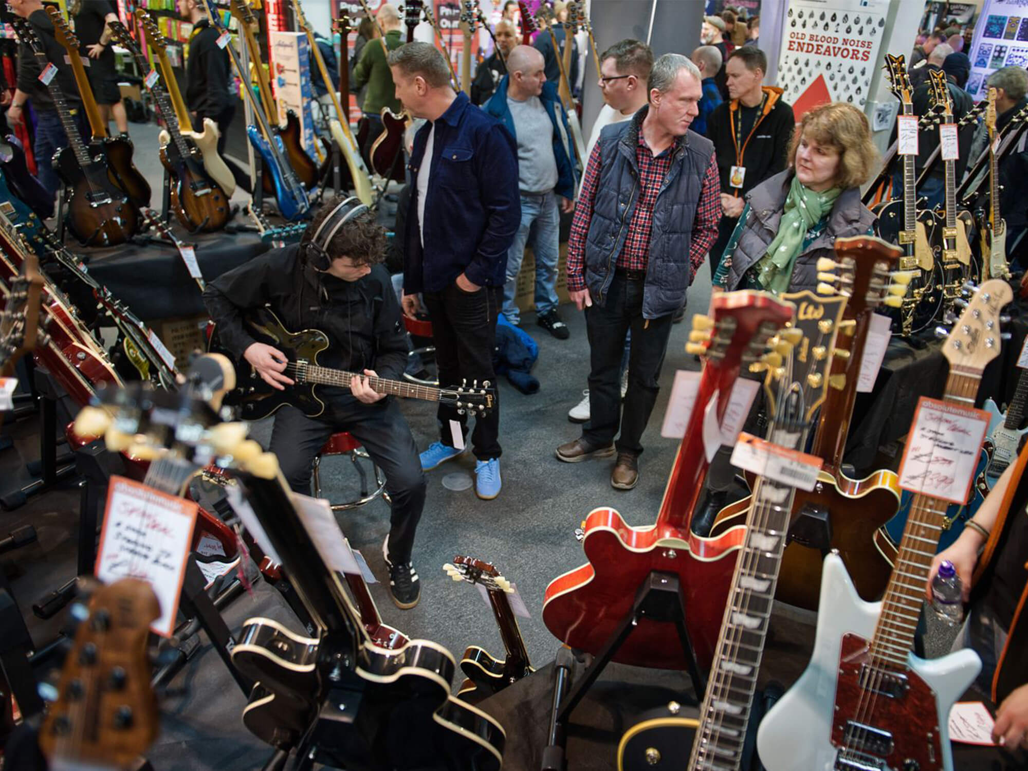 Exhibition at The Guitar show. There is a young person sat playing a guitar, with other people walking around near by. Lots of guitars are displayed on stands.