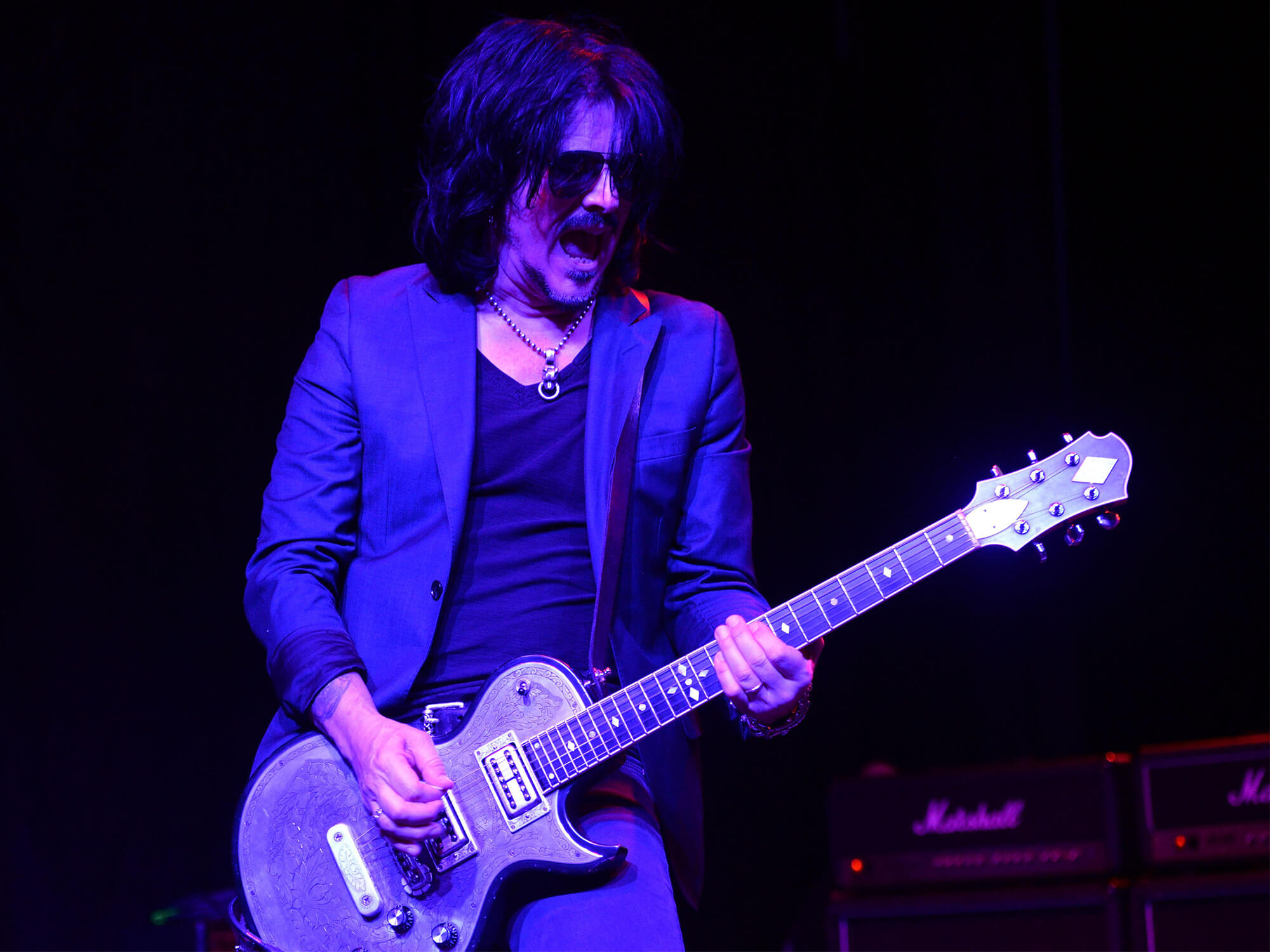 Gilby Clarke playing guitar under purple lighting. He's wearing shades and has his mouth open as if singing.