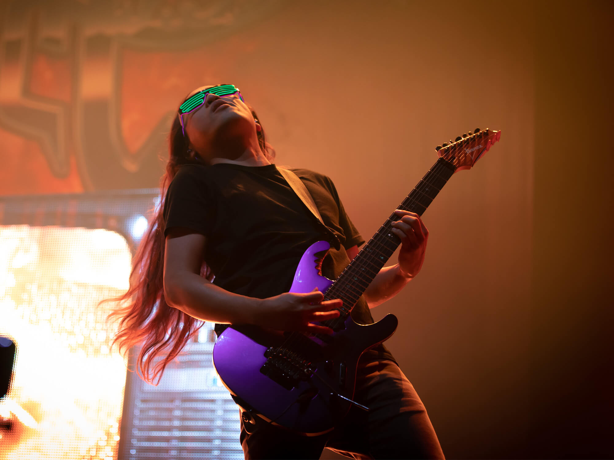 Herman Li on stage. He is playing a bright purple guitar and is wearing illuminating green shades.