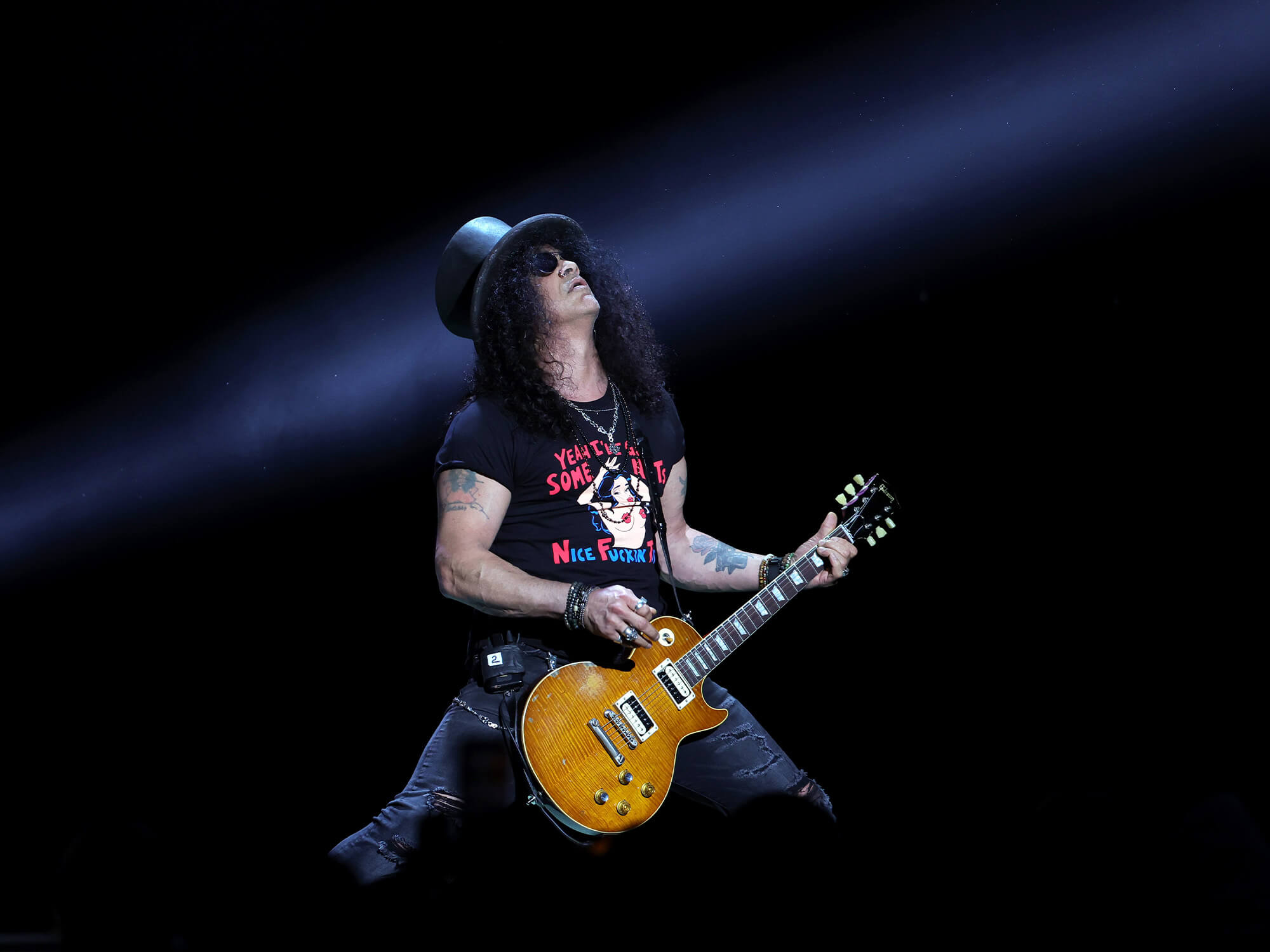 Slash playing his Les Paul on stage. He is wearing his famous top hat and is looking up.