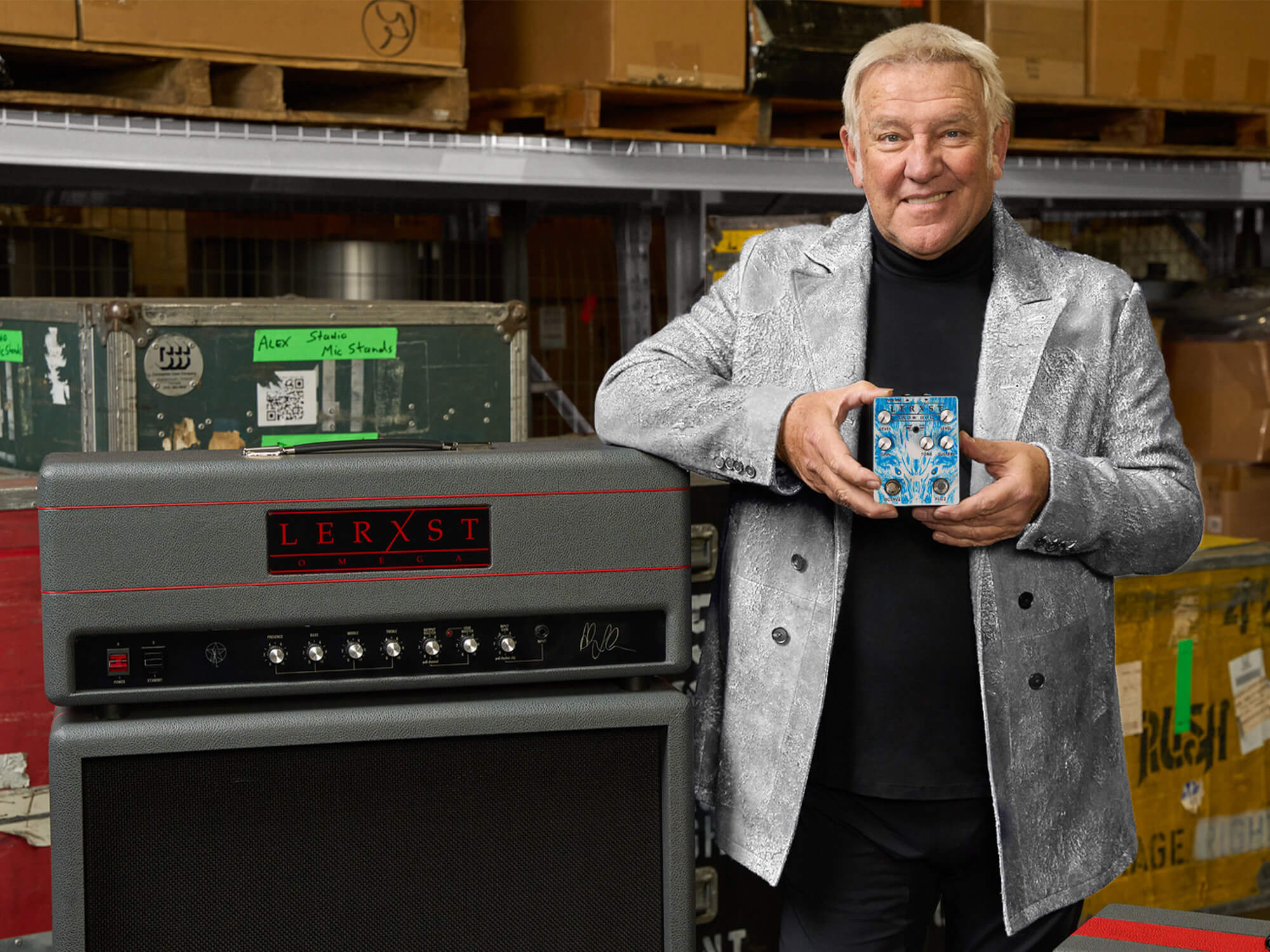 Alex Lifeson holding the Snow Dog pedal. He is smiling and stood next to a Lerxst amp stack.