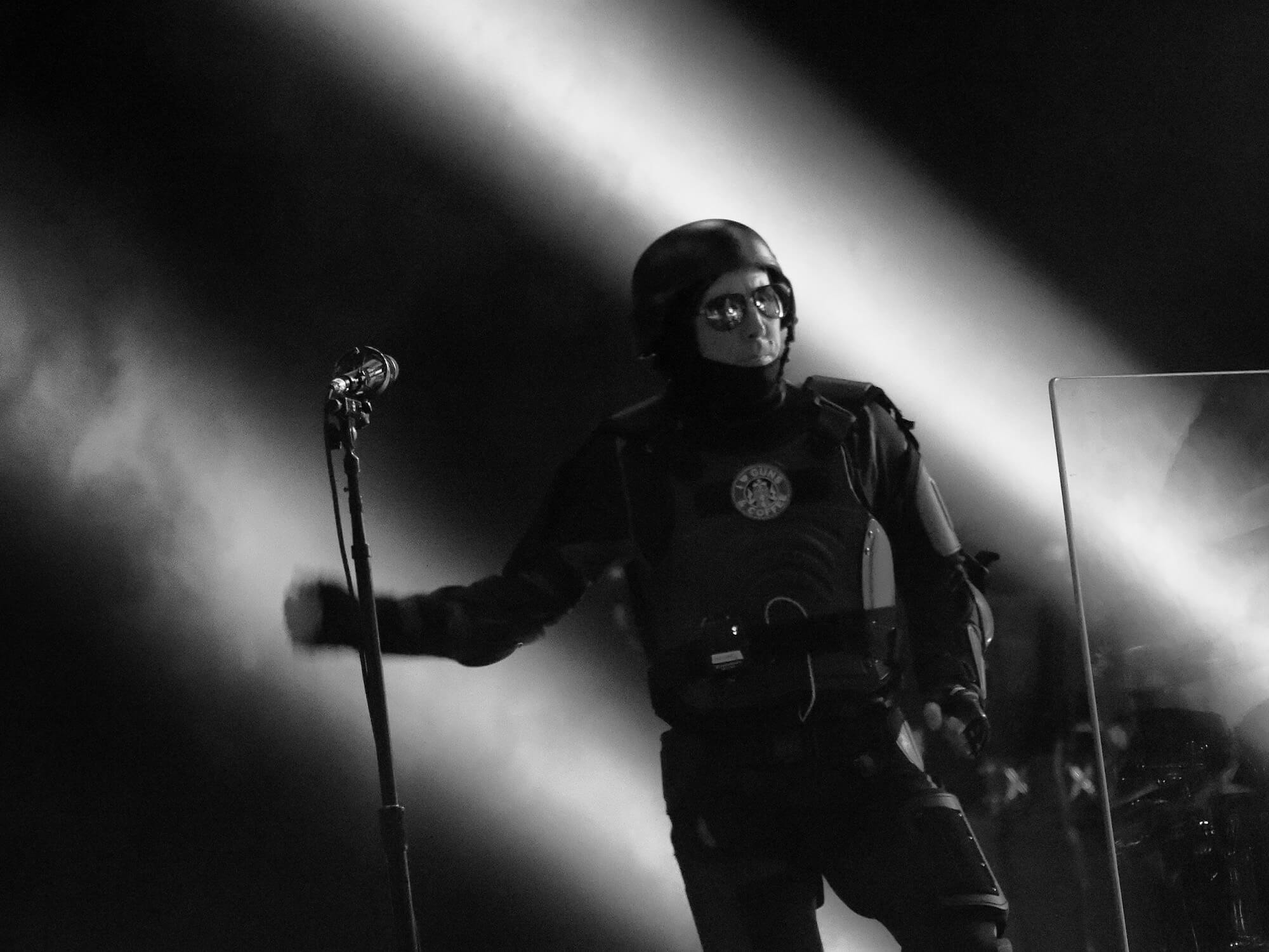 Maynard James Keenan on stage in 2017. He is wearing a helmet and combat suit and is stood next to a mic stand.