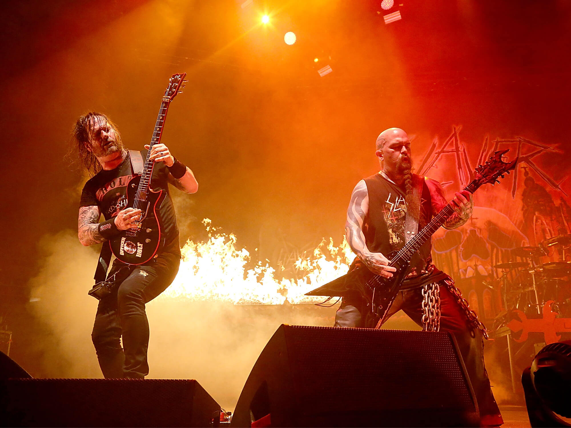 Gary Holt and Kerry King on stage in 2015. They're both playing guitar and standing in front of a flaming background.