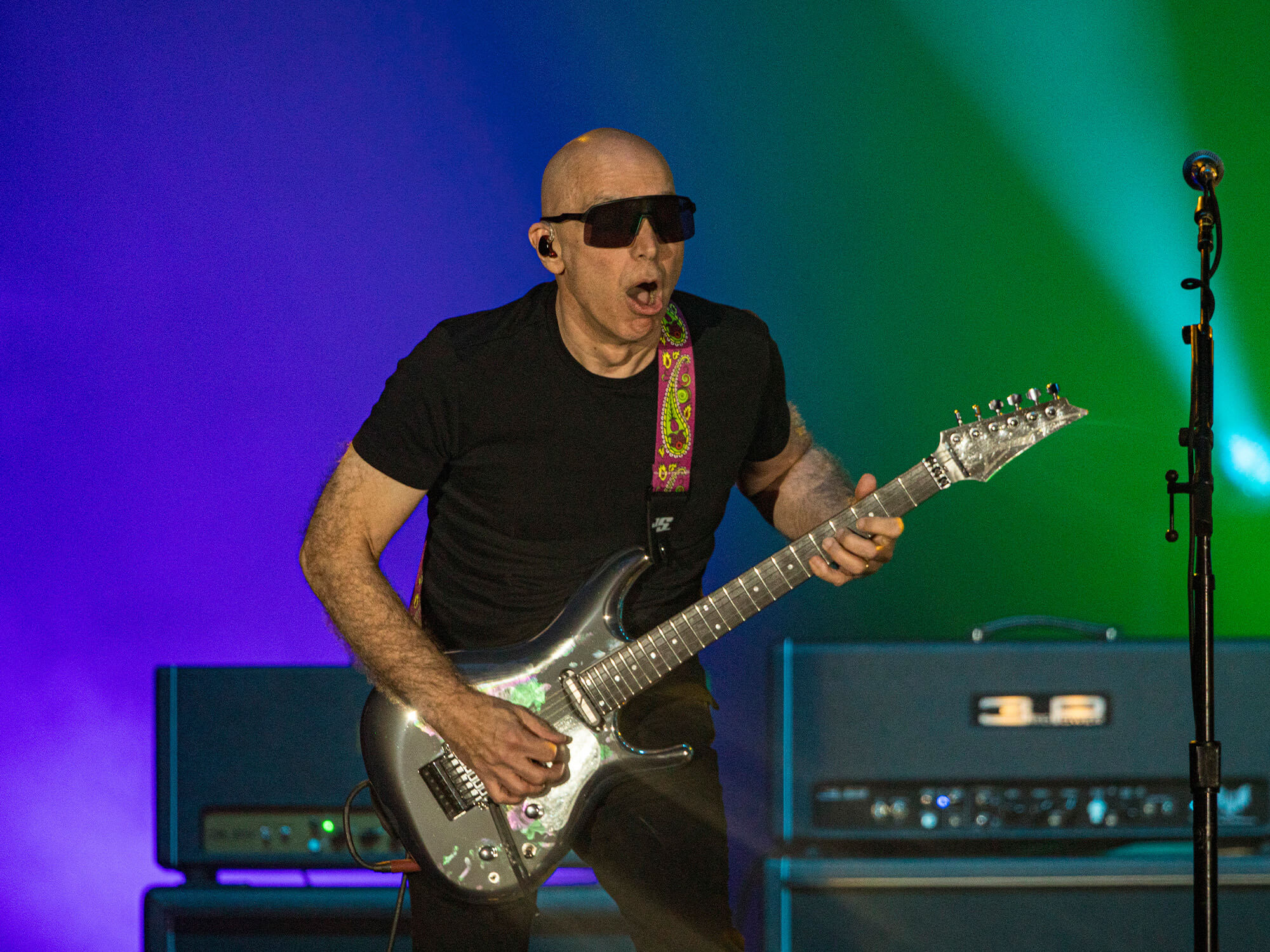 Joe Satriani playing guitar on stage. He is wearing his famed shades and has a shocked expression on his face.