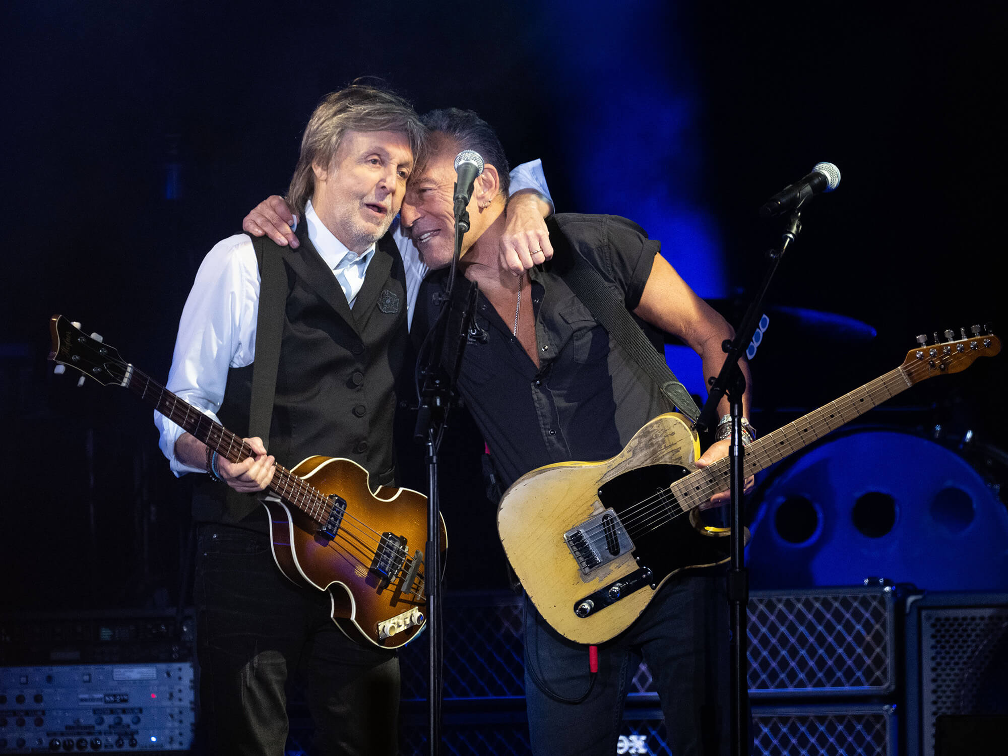 Paul McCartney and Bruce Springsteen on stage at Glastonbury in 2022. The pair each have one arm around the other and are holding their guitars in one hand.