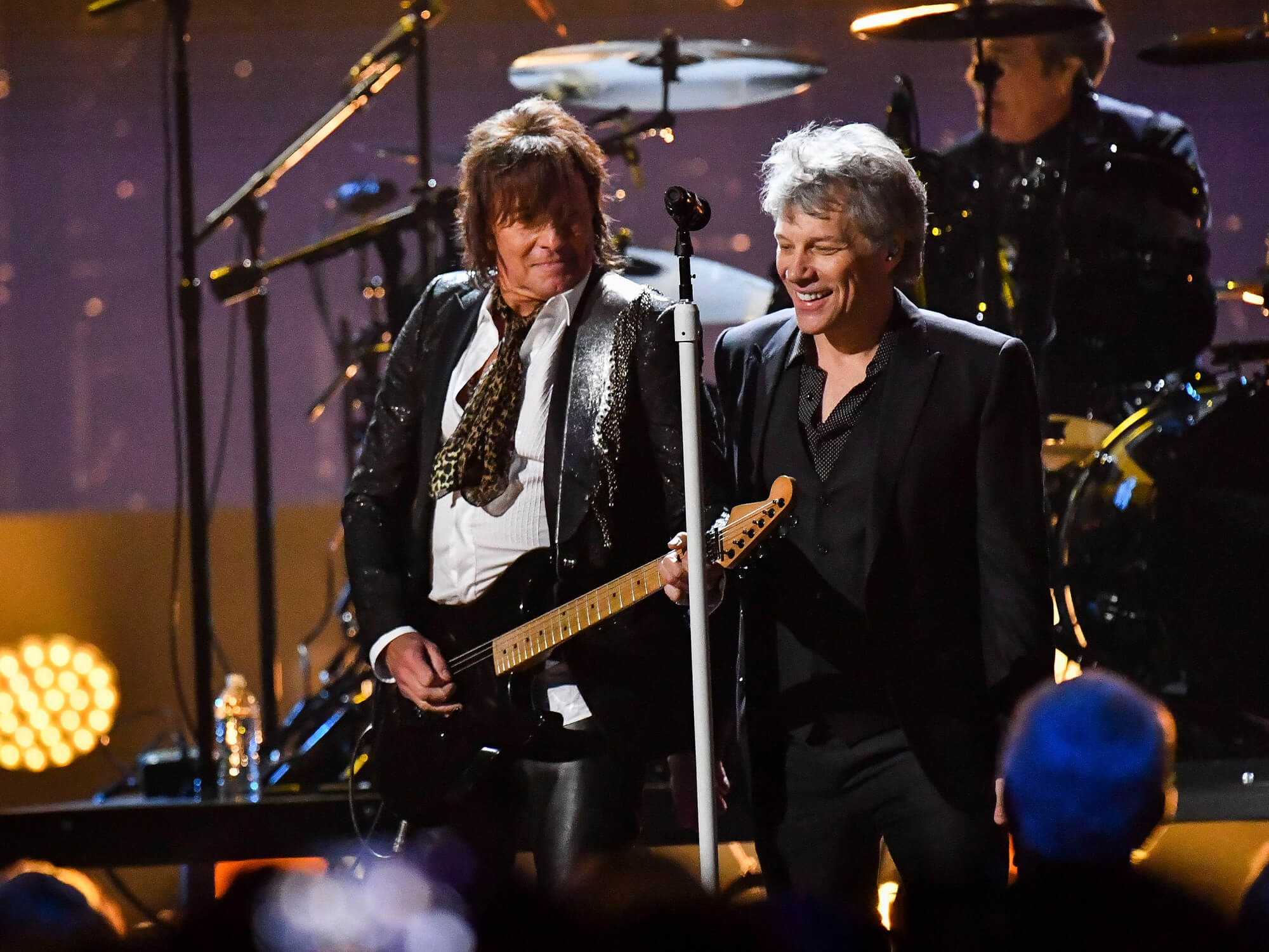 Richie Sambora and Jon Bon Jovi on stage. Richie has his guitar in hand, and Jon is standing by a mic stand and smiling.