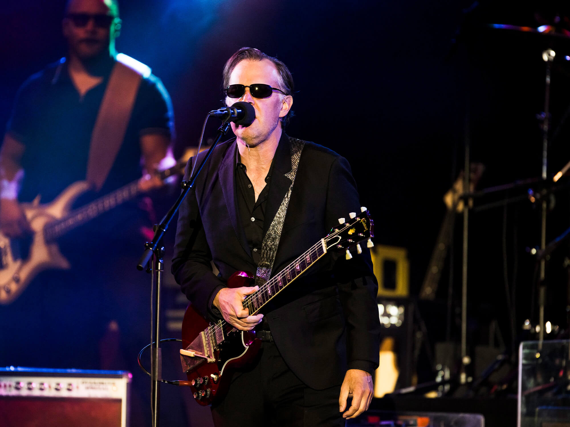 Joe Bonamassa in his classic sunglasses, holding the neck of a guitar as he is singing into a mic